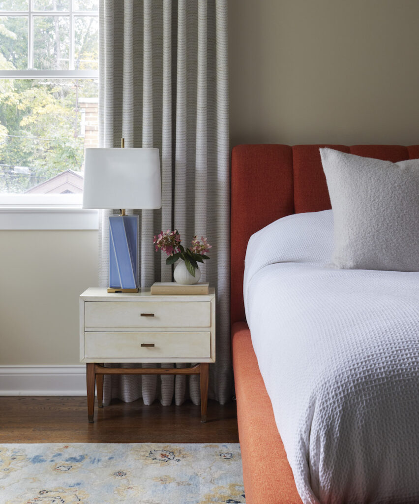 A bedroom vignette by Amy Kartheiser Design featuring a deep red headboard complementing the neutral bedding and accent decor in the space