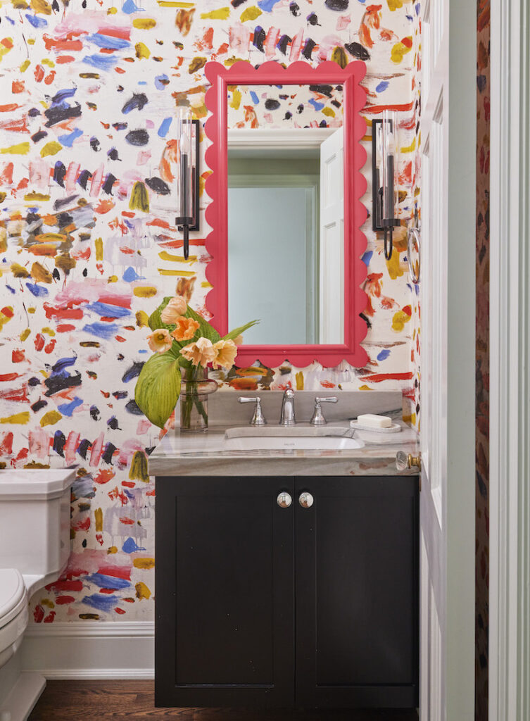 Vibrant small powder room with splashy abstract wallpaper, featuring a hot pink vintage framed mirror, a black vanity adorned with jewel handles, and contemporary candle sconces enhancing the overall look