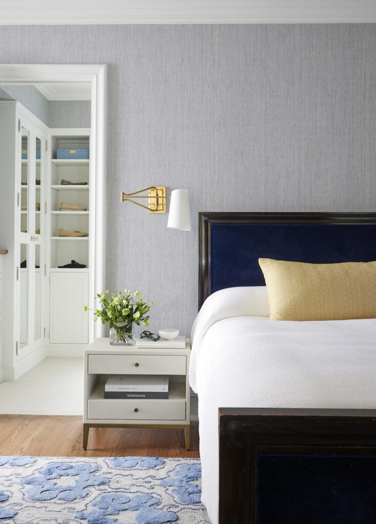 Bedroom vignette by a Chicago interior designer featuring a navy blue headboard paired with white bedding and pale yellow pillows. The setup includes a nightstand with accent decor, a contemporary sconce, and a walk-in closet displaying beautiful shoes, completing the composition