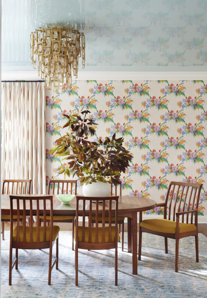 Colorful interior design: Stunning dining area featuring a light blue ceiling complemented by a contemporary tiered chandelier adorned with gold gilded accents and crystals. The walls are adorned with a floral-patterned wallpaper against a beige background, serving as a backdrop for a simple yet elegant wooden dining set