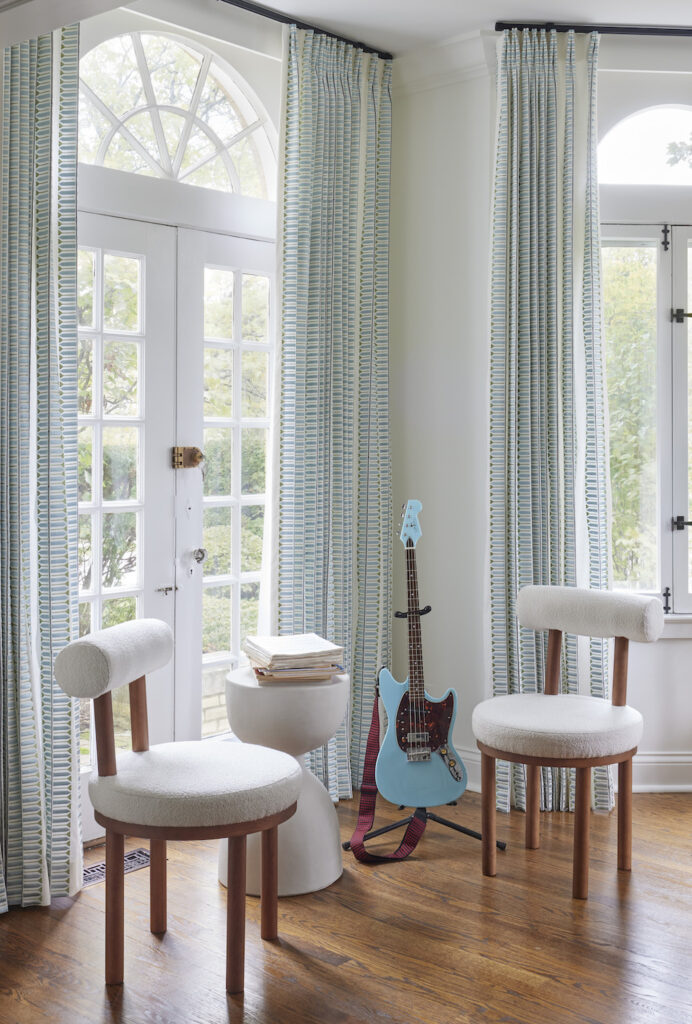 Living area corner featuring contemporary armless chairs set against large arched windows. Complemented by a sculptural side table, long aqua blue and white patterned drapes, and an electric blue guitar serving as an eye-catching accent