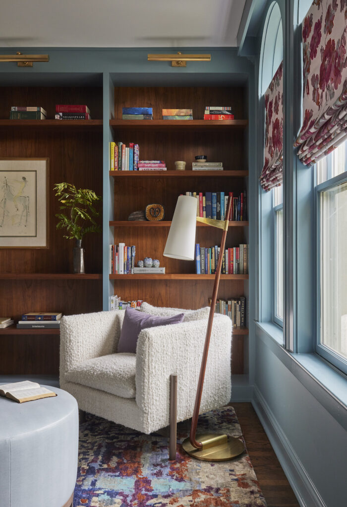 Maximalist reading room corner featuring a beige armchair with a purple cushion, set against light blue walls adorned with red and pink floral cafe curtains. A vibrant rug contrasts the armchair while harmonizing with the window hues. The composition is complemented by a dark wood bookshelf curated with colorful books and accent decor.