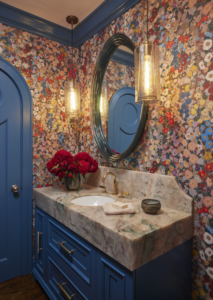 Vibrant and colorful powder room featuring a striking blue arched door and a maximalist floral wallpaper. The space is accentuated by a large round mirror, gold gilded hardware, hanging pendants, and a uniquely stained marble vanity top