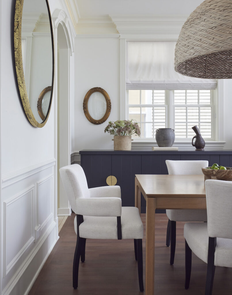 A beautiful dining area predominantly furnished in neutral tones, highlighted by a navy console, accent decor, and a woven bamboo pendant light above the dining space. The room is brought to life by a patina-layered antique round mirror