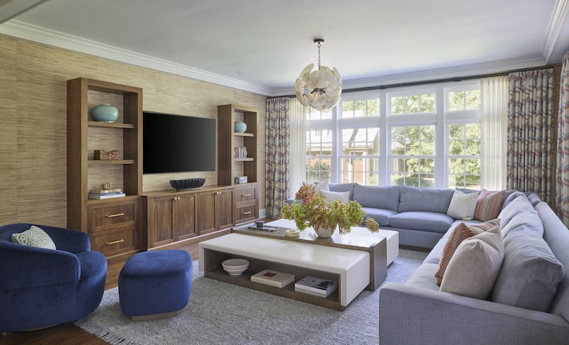 Curated interior design: An entertainment room featuring carefully chosen furniture pieces—a light blue sectional sofa, a contemporary coffee table with an inbuilt bookcase, a deep blue swivel chair with a footrest, a shell-inspired chandelier, and a classic wooden TV stand—all complemented by expansive windows and double drapes that flood the space with ample sunlight