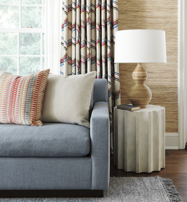 Light blue sectional sofa with beige and pastel-colored cushions, accompanied by a sculptural side table and lamp, complemented by beautifully patterned drapes in a colorful interior design vignette