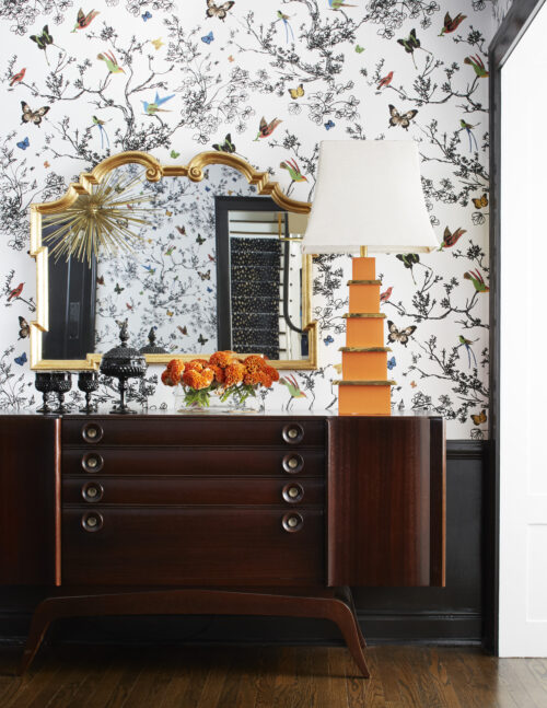 Floral wallpaper and black trim surround a dark wooden console, orange lamp, flowers, and mirror in a living space designed by Amy Kartheiser Design