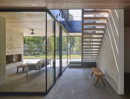 Floor-to-ceiling windows separating wooden staircase and outdoor living room with fireplace