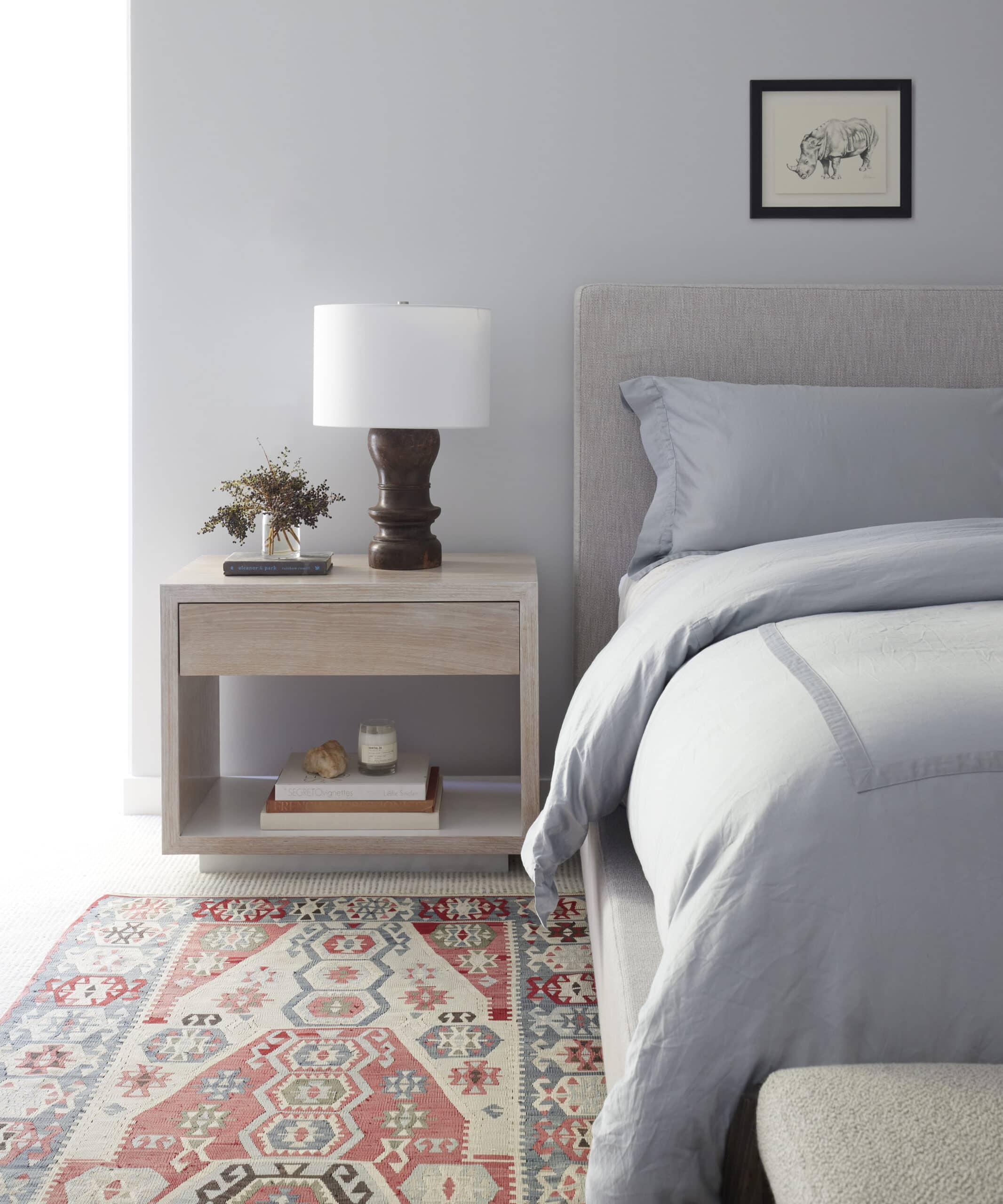 White bedroom with blue bedding, a wooden nightstand, a printed rug, and accessories.