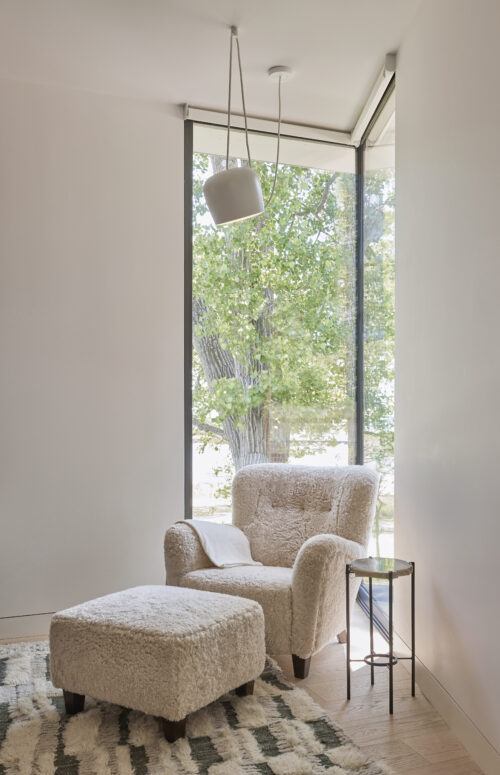 A corner reading nook featuring floor-to-ceiling windows, a boucle chair and ottoman, a side table, and rug.